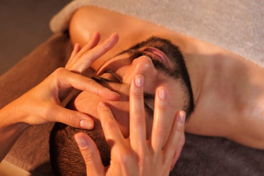 Close-up of female massage therapist's hands massaging man's forehead on dark background, effective facial massage for wrinkles, rejuvenation