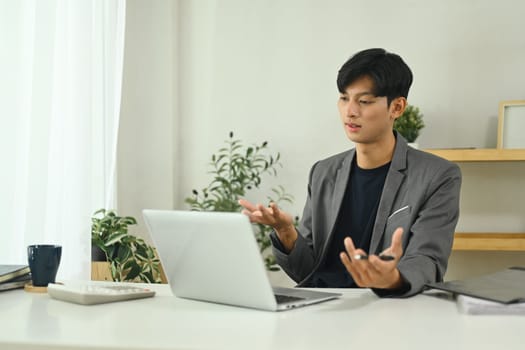 Young businessman in suit sitting at his workplace in front of laptop and communicating by video call.