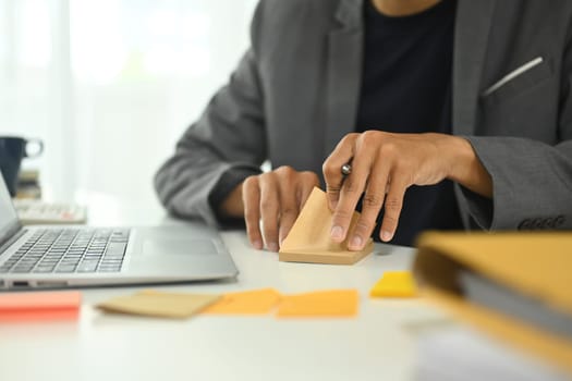 Unrecognizable sitting at a desk with a laptop and writing idea on adhesive note.