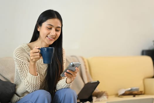 Happy young woman sipping coffee and checking message on her mobile phone.