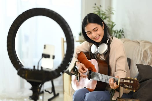 Smiling female music influencer recording video tutorial on playing guitar.