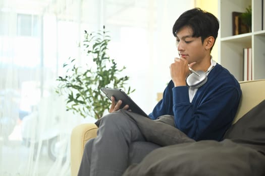 Thoughtful young male freelancer using digital tablet while sitting on couch.