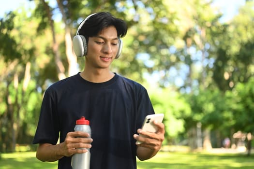 Young athletic man taking a break outdoors, drinking water and using mobile phone.