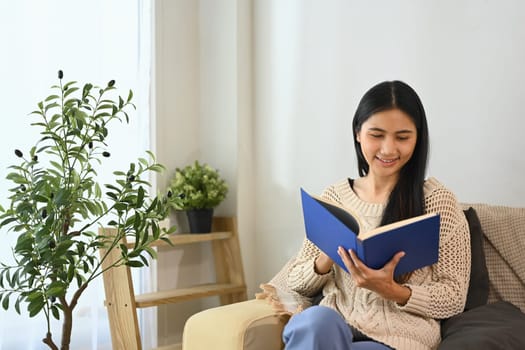 Smiling young woman reading book on sofa at home.