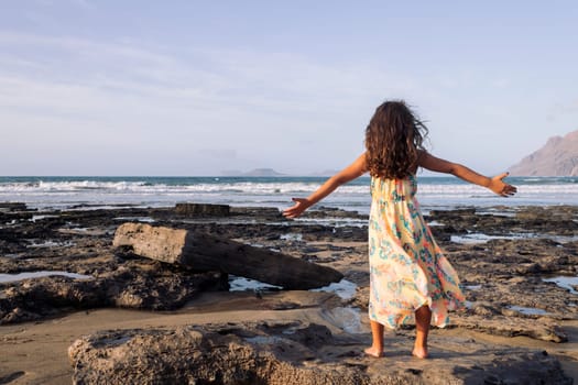 rear view of unrecognizable little girl in summer dress with open arms contemplating sea from Famara Beach in Lanzarote, Canary Islands, Spain, concept of travel and wild nature