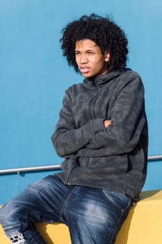 serious african american teenager with curly hair sitting on a yellow wall with arms crossed in front of blue background, concept of adolescence and young lifestyle