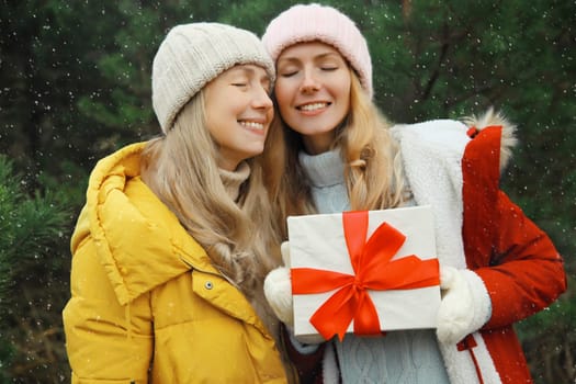 Happy smiling women friends holding festive gift box, celebration, joyful girlfriends together on Christmas tree background in winter forest, wearing hat, jacket outdoors