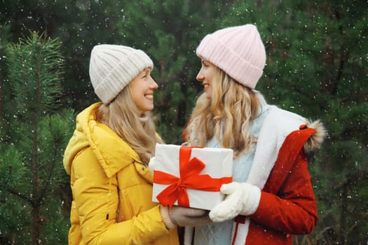 Happy smiling women friends holding festive gift box, celebration, joyful girlfriends together on Christmas tree background in winter forest, wearing hat, jacket outdoors