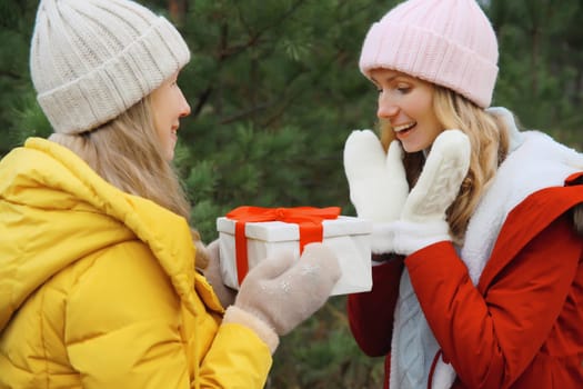Happy smiling women friends gives festive gift box, celebration, joyful girlfriends together on Christmas tree background in winter forest, wearing hat, colorful jacket outdoors