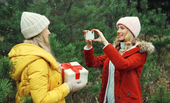Happy smiling women friends taking picture with phone, festive gift box, celebration, joyful girlfriends together on Christmas tree background walk in winter forest, wearing hat, jacket outdoors