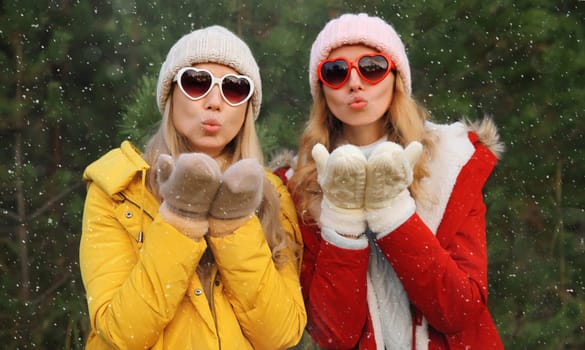Portrait of happy women friends having fun on Christmas tree background in winter forest, celebration, joyful girlfriends blowing kiss together in heart shaped glasses, wearing hat, jacket outdoors