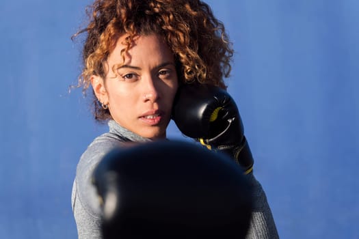 determined adult female boxer with curly hair ready to fight on boxing gloves during workout on blue background, concept of strength and healthy lifestyle