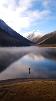 A lone angler stands fishing in a serene mountain valley, with snow capped peaks and misty waters.