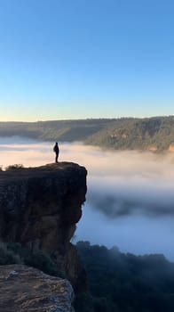 A lone person stands on a cliff edge, gazing over a sea of clouds with a stunning view of the forested hills.