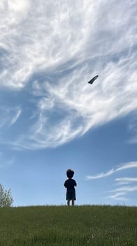 A young child standing on a grassy hill, flying a kite under a sky filled with wispy clouds.