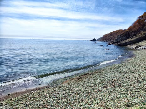 Waves gently lap against a pebbly beach under a clear blue sky near rocky cliffs.