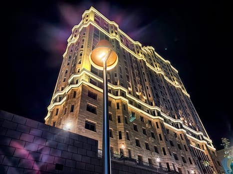 The building stands tall against the nighttime sky, illuminated by warm lights outlining its architectural features, reflecting vibrant colors.
