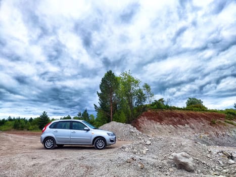 A silver vehicle stands on rocky ground surrounded by greenery and a stunning cloud-filled sky on a sunny day.