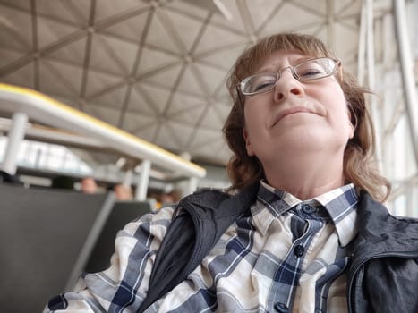 A weary middle-aged woman waits in an airport terminal, showing signs of fatigue as she anticipates her delayed flight departure amid the bustling crowd.