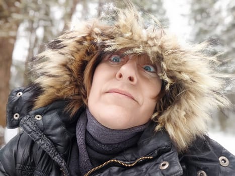 Funny middle aged woman in a winter coat with fur, scarf taking selfie on nature outdoors and cold, snowy background with blue sky and white clouds