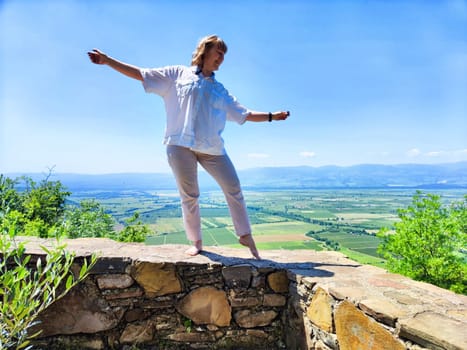 A woman enjoys a moment of balance atop a stone wall, overlooking expansive green valleys and mountains on a sunny day.