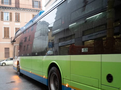 Cremona, Lombardy, Italy October 15th 2024 Green electric bus driving on wet city street reflecting buildings on its side in a rainy day