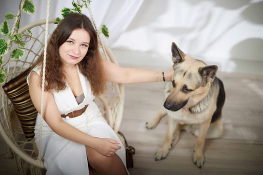 A young woman relaxes in a swing while her German Shepherd rests peacefully beside her in a bright, airy room.