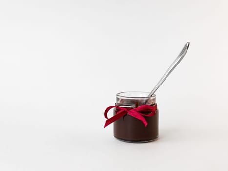 Small glass jar of chocolate spread with red ribbon and spoon on white background.