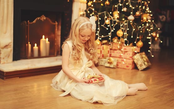 Happy little girl child in dress with festive gift box sitting near Christmas tree and fireplace with holiday lights at home