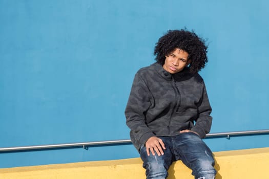 serious african american teenager with curly hair sitting on a yellow wall looking to the side in front of blue background, concept of adolescence and young lifestyle, copy space for text