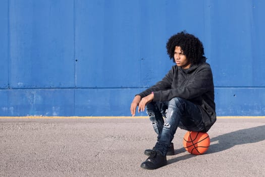 serious african american teen male in informal outfit sitting on basketball ball near blue background and looking to the side, concept of sport and young lifestyle, copy space for text