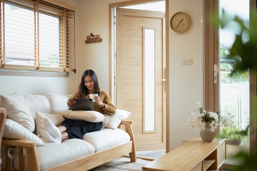 Relaxed young woman drinking hot tea and using digital tablet in living room.