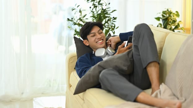 Cheerful young man using smartphone and relaxing on sofa.