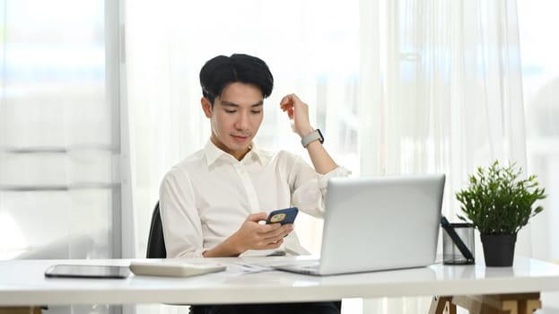 Smiling young entrepreneur in white shirt sitting in office and using smartphone.