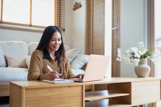 Charming young woman working remotely with laptop and writing notes.