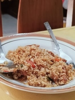 A plate of flavorful fried rice is served with a spoon and a fork on a wooden table. The dish includes pieces of vegetables and meats, showcasing a homemade and appetizing meal.