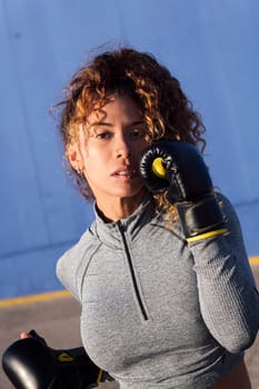determined adult female boxer with curly hair ready to fight on boxing gloves during workout on blue background, concept of strength and healthy lifestyle