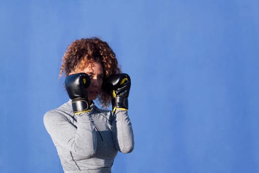 determined adult female boxer with curly hair ready to fight on boxing gloves during workout on blue background, concept of strength and healthy lifestyle, copyspace for text