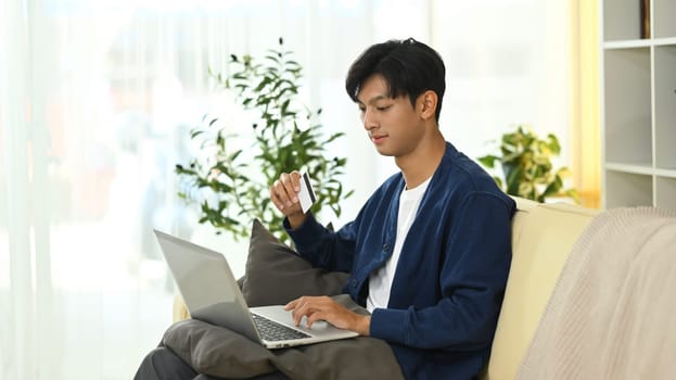 Handsome asian man holding credit card making purchase via laptop.
