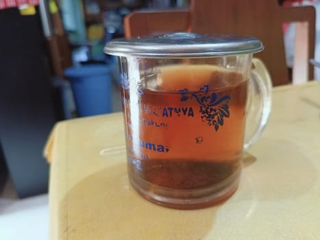 Warm glass of tea with a floral design on a clear mug with metal lid, placed on a wooden table. Perfect for cozy moments and relaxation. Captures a traditional tea-drinking experience.