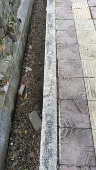 An urban scene showing the contrast between a patterned sidewalk and a dirt pathway. The textured surfaces highlight urban decay and natural elements coexisting in a city environment.