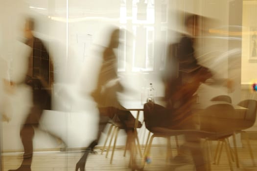 A blurry image of people walking in a room with chairs and a table. The people are wearing business attire and appear to be in a hurry. The chairs and table are arranged in a semi-circle
