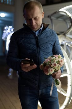 A man in a blue jacket and jeans walks through a city at night. He is looking down at his phone in his right hand and holding a bouquet of pink and white flowers in his left hand.