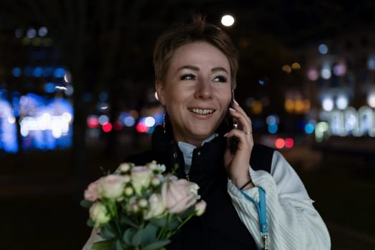 A woman with short hair is walking through a city at night. She is wearing a dark jacket and a white shirt. She is talking on her phone and holding a bouquet of white and pink flowers.