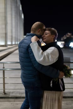 A man and a woman are hugging in the city at night. The man is wearing a blue jacket and jeans, and the woman is wearing a white shirt, a black vest, and a beige skirt.