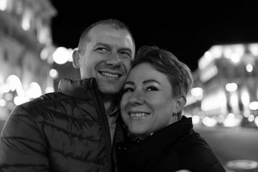 A man and a woman, presumably a married couple, are walking together in a city at night. They are both smiling and looking at the camera.
