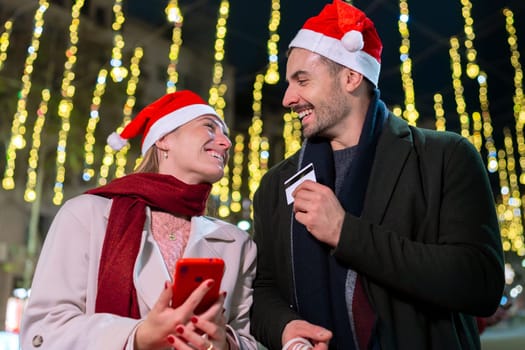 Smiling couple wearing Santa hats, standing under festive Christmas lights. They look at each other warmly, creating a joyful holiday atmosphere. One holds a phone, capturing memories.