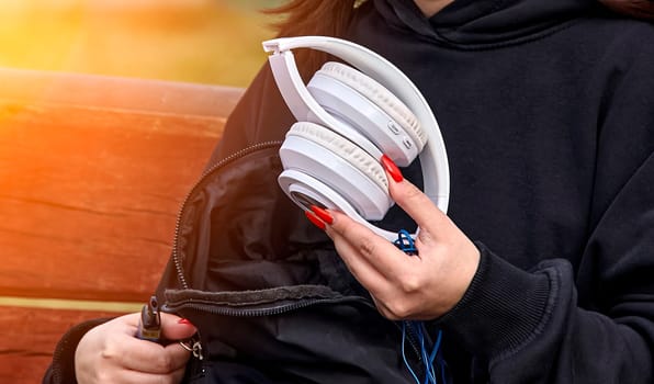 A woman wearing a black hoodie sitting on a bench in a sunny park, holding white over-the-ear headphones with a cord. The background is blurry, creating a serene outdoor setting.