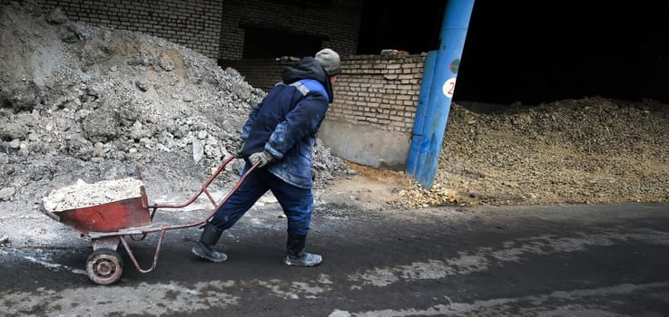 A man in a blue jacket and work boots pushes a wheelbarrow filled with gravel along a dirt path. The path leads to a brick wall and a pile of gravel.