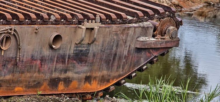 A close-up of a rusted dredge, its treads and hull resting on the bank of a tranquil waterway. The stillness of the water reflects the surrounding landscape.